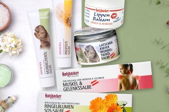 Eine Sammlung von natürlichen Hautpflegeprodukten, darunter Cremes, Salben und Öle, die auf einem grünen Hintergrund mit Blumen und Kräutern ausgestellt sind. Zu den Produkten gehören lavendelduftende Artikel, Lippenbalsam und eine Gelenk- und Muskelpflegesalbe.