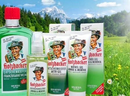 Ein Bild zeigt verschiedene Flaschen und Tuben von Franzbranntwein Produkten in einem grünen und weißen Farbschema, vor dem Hintergrund einer grünen, üppigen Wiese mit Bergen und Sonnenschein.