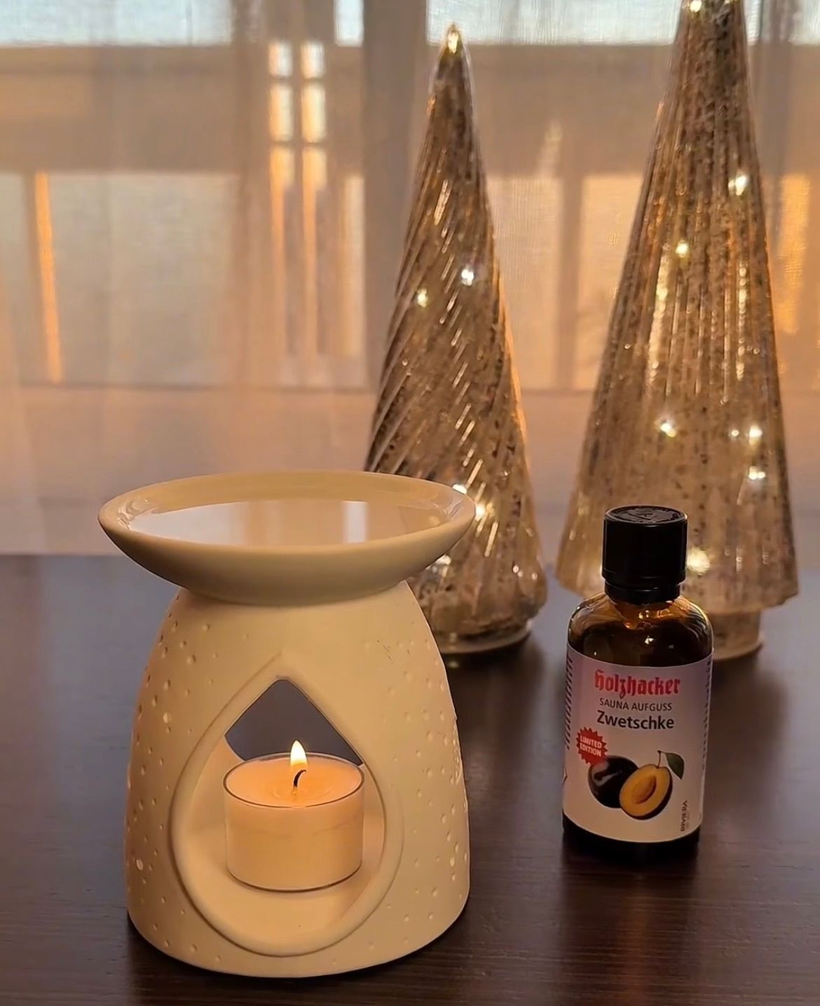 Eine Kerze brennt in einem Keramik-Kerzenhalter auf einem Holztisch, mit zwei Glas-Weihnachtsbäumen im Hintergrund. Neben dem Kerzenhalter steht eine Flasche ätherisches Öl.