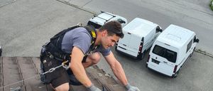 Ein Mann ist auf einem Dach und repariert Dachziegel, während er einen Sicherheitsgurt trägt. Hinter ihm sind mehrere weiße Lieferwagen auf der Straße und ein Wohnwagen geparkt.