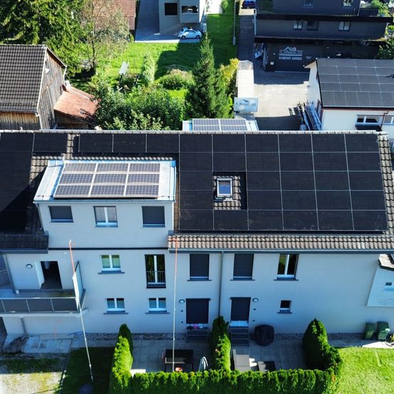 Eine Vogelperspektive eines Wohngebäudes mit Solarmodulen auf dem Dach. Bäume und andere Häuser sind im Hintergrund sichtbar.