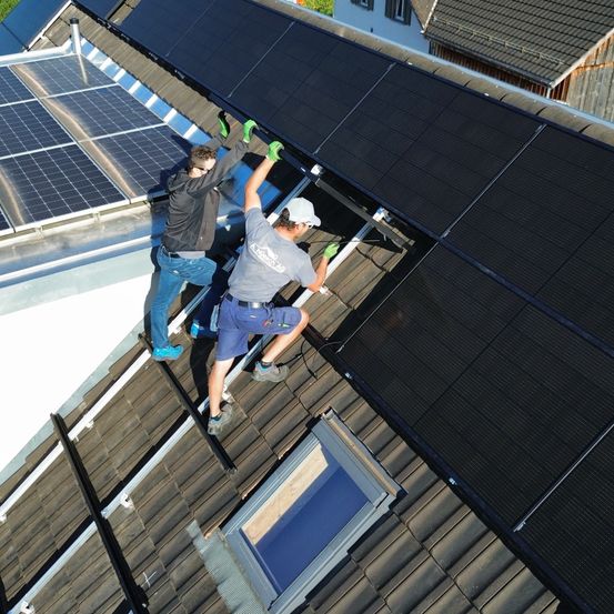Zwei Arbeiter installieren Solarpanele auf einem Dach. Einer trägt ein graues Shirt und eine weiße Kappe, der andere eine schwarze Jacke und blaue Jeans. Solarpanele bedecken das Dach, mit einem Fenster im Vordergrund sichtbar.