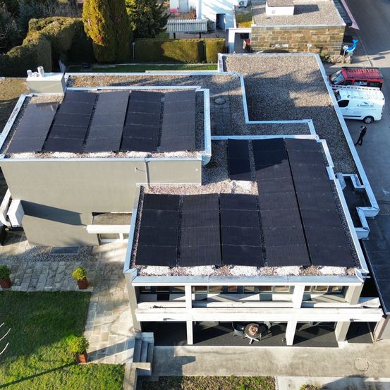 Ein Luftbild eines modernen Hauses mit einem Schotterdach und installierten Solarmodulen. Das Haus ist von Gras und Büschen umgeben. Eine Person geht in der Nähe der Straße.