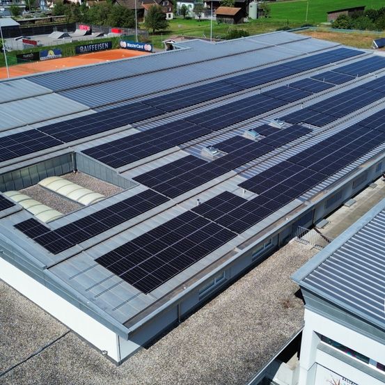 Eine Draufsicht auf ein großes Gebäude mit zahlreichen Solarpanelen auf dem Dach. Das Gebäude hat eine graue Außenfassade und die Solarpanele sind auf einem flachen Dach montiert.