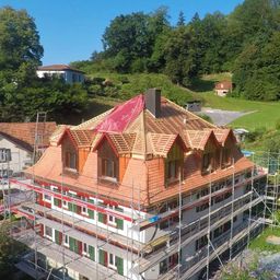 Ein Haus im Bau mit einem roten Dach, umgeben von Gerüsten, in einer ländlichen Gegend mit grünen Feldern und Bäumen.