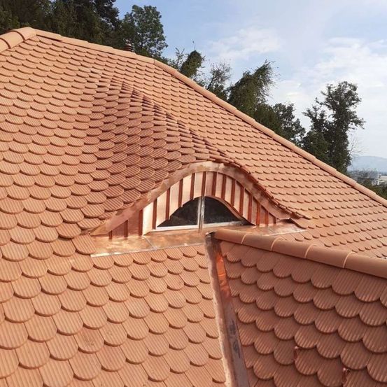 Ein Dach mit Terrakotta-Ziegeln und einem Fenster in einem Kupferrahmen. Das Dach hat ein wellenförmiges Muster und ist von grünen Bäumen umgeben.