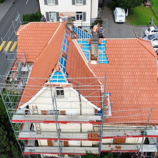 Luftaufnahme eines Hauses in Renovierung mit gefliestem Dach, Gerüst und Arbeitern. Zwei Arbeiter sind auf dem Dach, einer ohne Oberteil, und ein weißer Van ist in der Nähe geparkt.