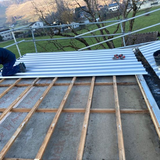 Ein Mann arbeitet auf einem Dach und installiert Metallbleche. Werkzeuge und Gerüste sind in der Nähe. Hinter ihm sind ein Baum und ein Haus zu sehen.