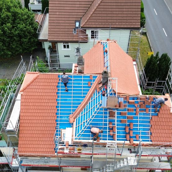 Luftaufnahme eines Hauses im Dachbau. Arbeiter installieren Ziegel auf dem Dach. Das Haus hat einen Schornstein, und es gibt Gerüste drum herum.