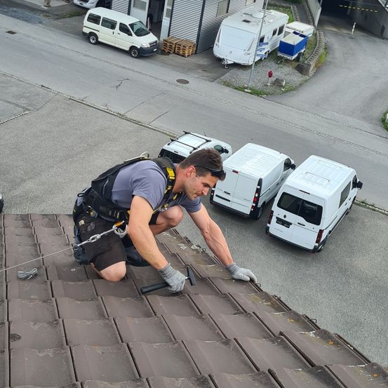 Ein Mann ist auf einem Dach, trägt einen Gurt und Handschuhe und hält einen Hammer. Mehrere weiße Lieferwagen sind am Straßenrand geparkt.