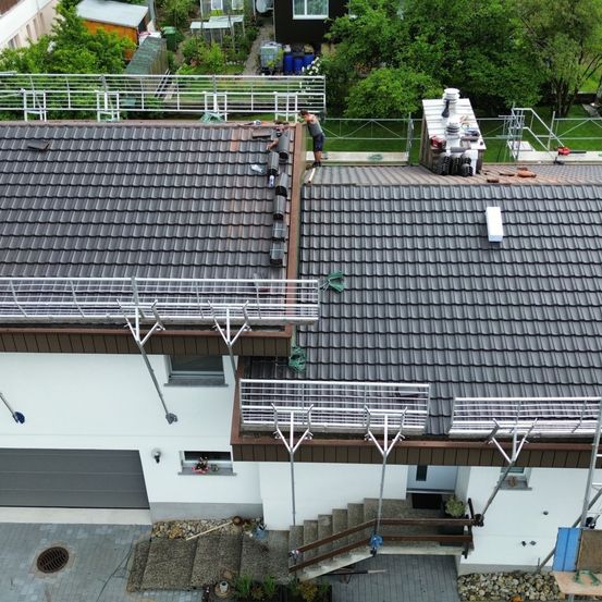 Eine Vogelperspektive eines Hauses mit repariertem Dach. Eine Person steht auf dem Dach, während eine andere auf der Gerüstarbeit ist. Das Haus hat eine weiße Außenseite und graue Dachdeckung.