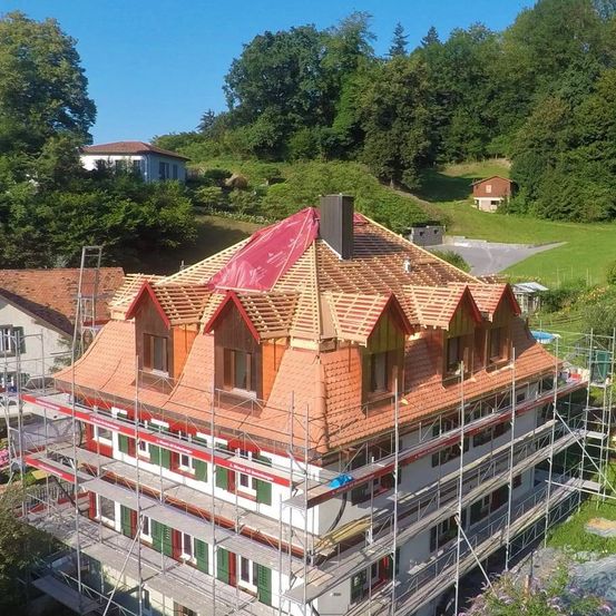 Ein Luftbild eines großen Hauses im Bau. Das Dach ist teilweise mit roten Ziegeln bedeckt und Gerüste umgeben das Gebäude. Im Hintergrund befinden sich Bäume und eine grüne Wiese.