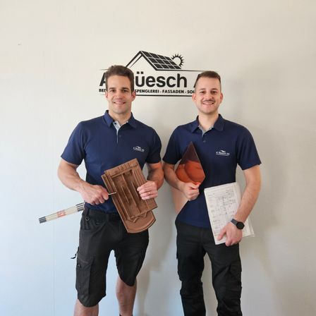 Zwei Männer in blauen Hemden und schwarzen Hosen stehen nebeneinander. Sie halten ein Holzobjekt und ein Stück Papier. Hinter ihnen ist eine weiße Wand mit einem Schild.