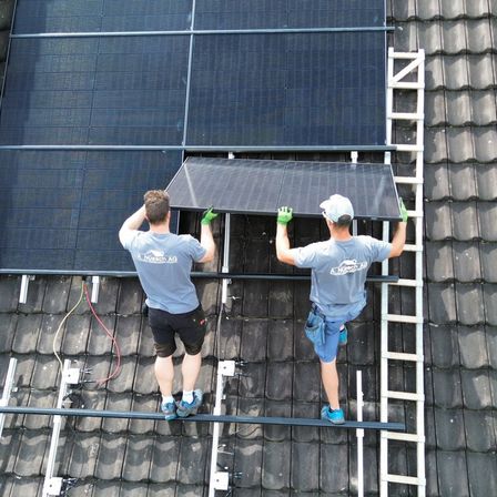 Zwei Männer installieren Solarpanele auf einem Dach. Ein Mann hält das Panel, während der andere auf einer Leiter steht. Sie tragen Handschuhe und blaue Shorts.