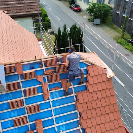 Ein Dachdecker installiert neue Ziegel auf einem Dach. Er trägt Kopfhörer und eine Sicherheitsgurt. Das Dach hat eine blaue Unterlage und rote Ziegel.