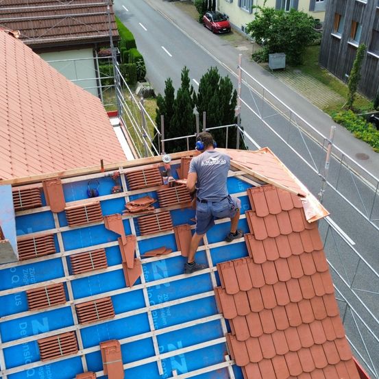Ein Dachdecker installiert neue Ziegel auf einem Dach. Er trägt Kopfhörer und eine Sicherheitsgurt. Das Dach hat eine blaue Unterlage und rote Ziegel.