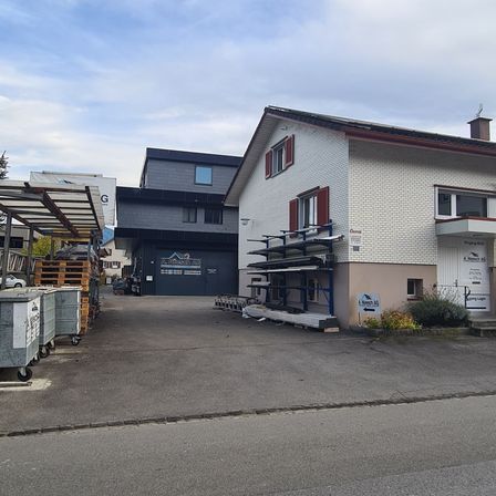 Ein Außenbereich mit zwei Gebäuden, von denen eines eine Werkstatt mit einer Garagentür ist. Lagerboxen und Materialien befinden sich in der Nähe der Werkstatt. Ein weißes Haus mit Schornstein steht daneben.