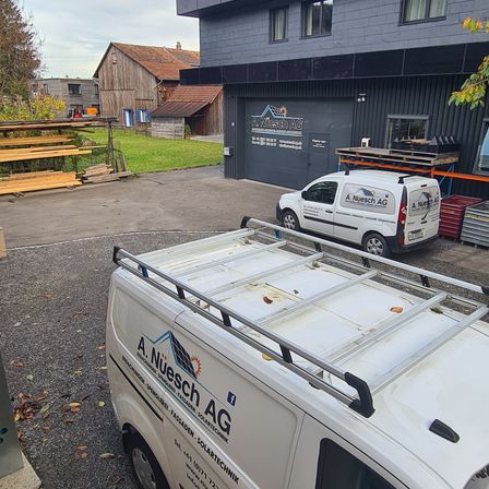 Ein weißer Lieferwagen mit Dachträgern ist vor einem Gebäude mit dem Schriftzug 'A. Nuesch AG' geparkt. Hinter dem Lieferwagen steht ein weiterer weißer Lieferwagen. Das Gebäude hat eine graue Wand und mehrere Fenster. Seitlich befinden sich Holzplanken und ein Metallpalette. In der Ferne steht ein Haus mit einem braunen Dach.