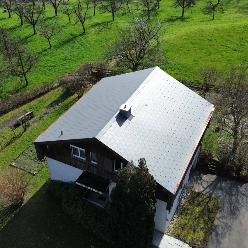 An aerial view of a house with a gray slate roof, surrounded by a green field and trees.