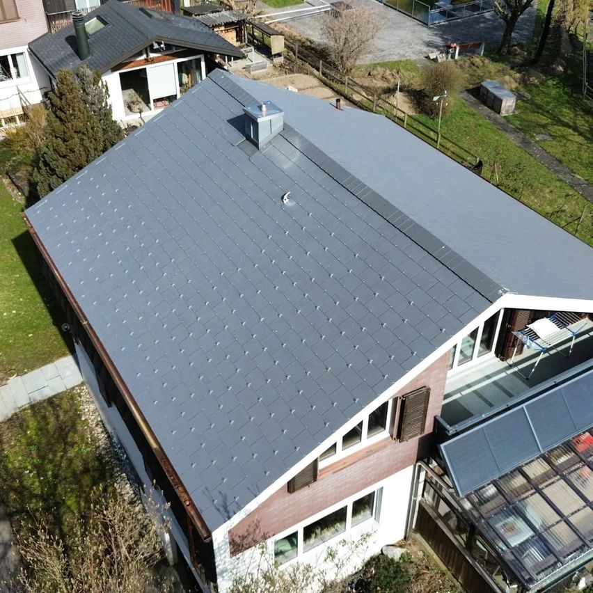 Aerial view of a modern house with a gray tiled roof and solar panels on the right side. The house has a brown wooden facade and white walls.