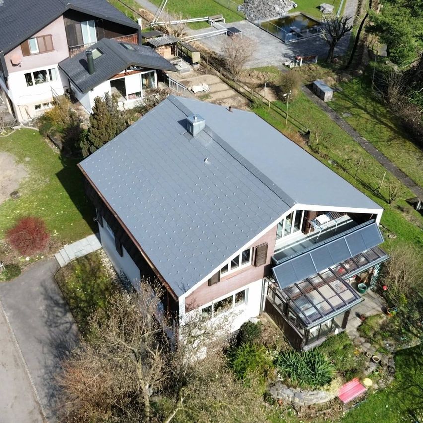 An aerial view of a modern house with a gray roof and solar panels, surrounded by greenery and a driveway.