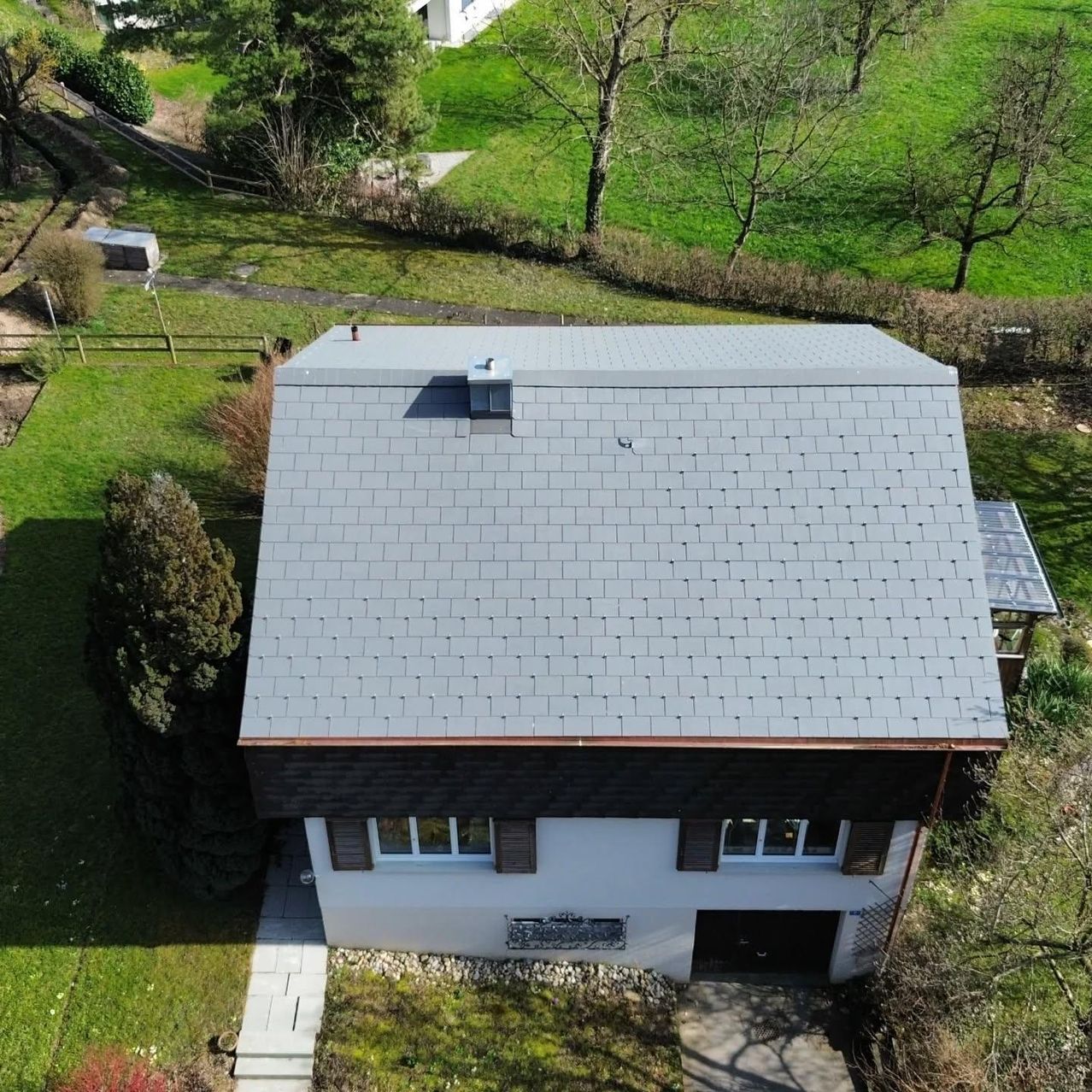 An aerial view of a house with a gray slate roof and a white exterior. The house has a chimney and several windows. It is surrounded by a green lawn and trees.
