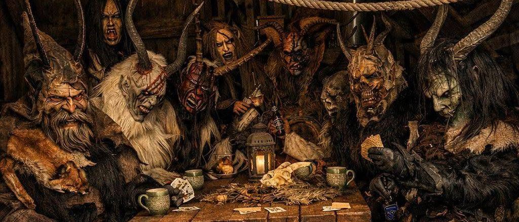 Eine Gruppe von Personen in Hörnermasken und Fellen sitzt um einen Holztisch herum, hält Tassen und Karten und wird von einer Laterne beleuchtet.