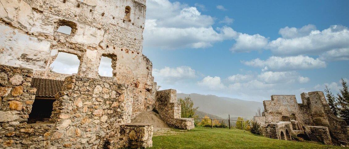 Eine Ruine einer Steinburg mit einem grasbewachsenen Hügel, blauem Himmel mit Wolken und entfernten Bergen unter einem klaren Himmel.
