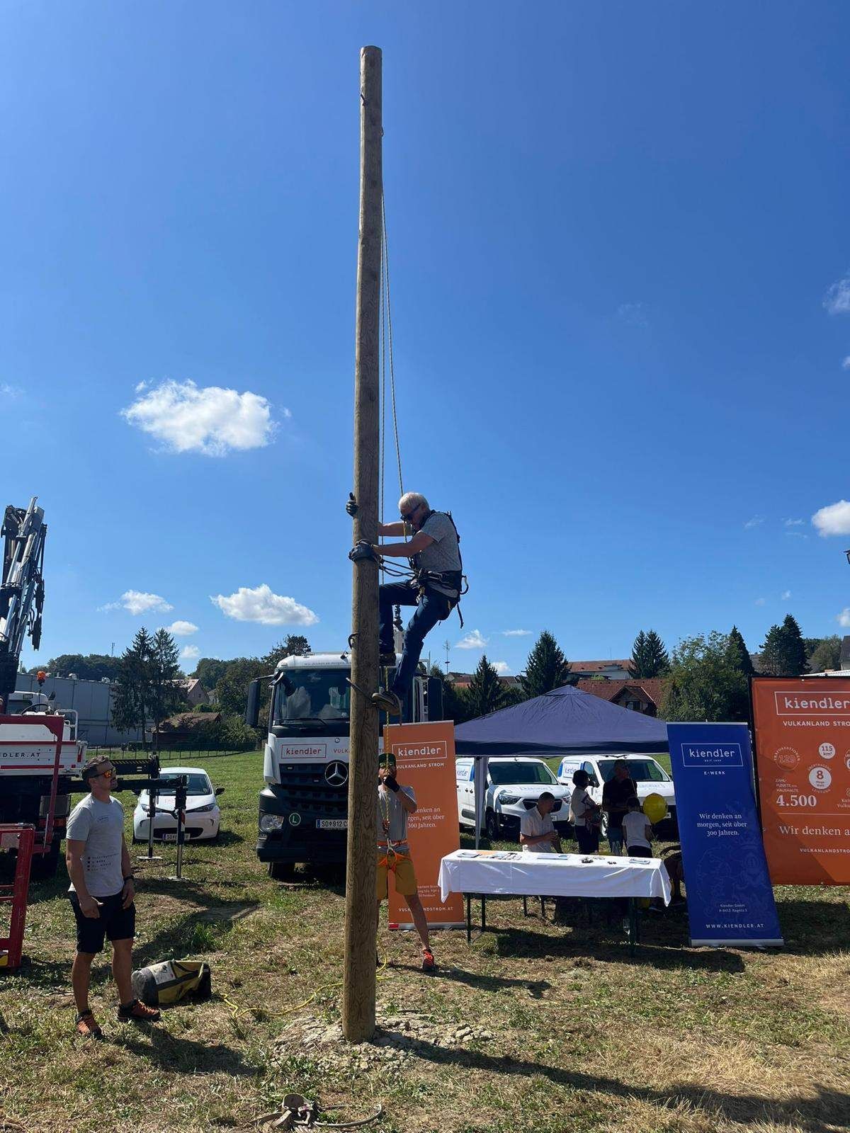 A man climbs a tall pole outdoors while a few people stand around, some with a banner. A tent and vehicles are nearby.