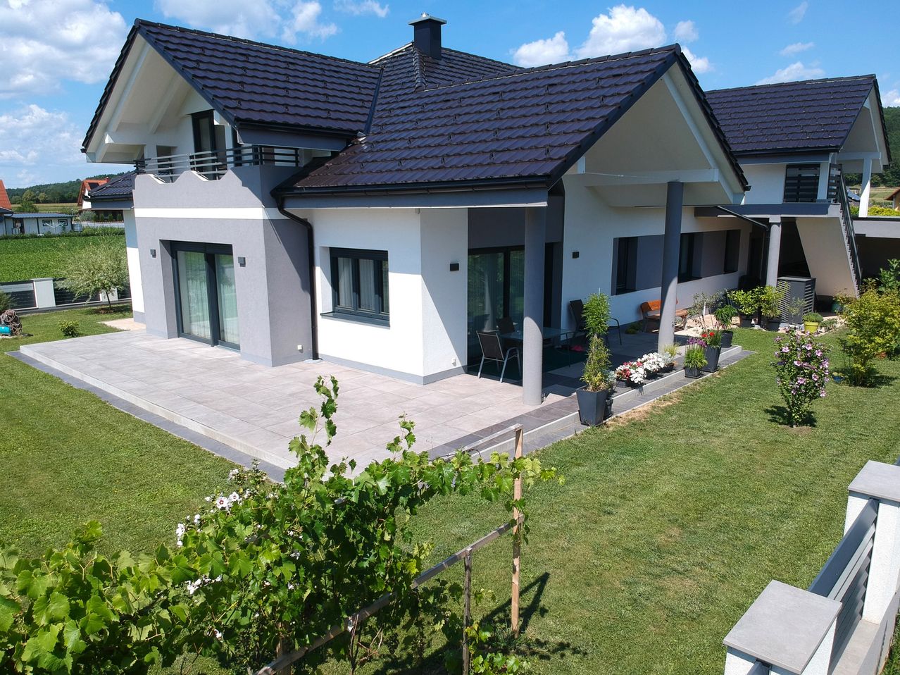 Ein modernes Haus mit einem gefliesten Dach und einer gepflasterten Terrasse. Topfpflanzen und Blumen schmücken den Vorgarten. Ein Balkon mit Geländern und Glastüren.