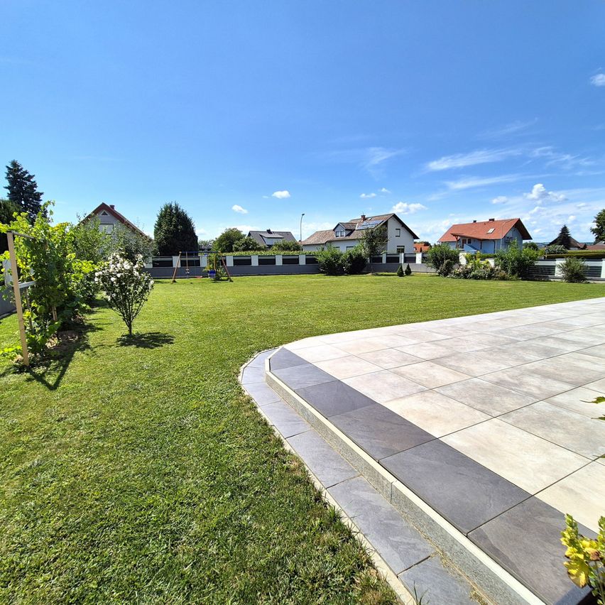 Ein breiter, gepflegter Rasenbereich mit einem gepflasterten Gehweg. Dahinter ein Haus mit rotem Dach, umgeben von Bäumen und anderen Häusern. Links ist ein Spielplatz zu sehen. Der Himmel ist blau mit einigen Wolken.