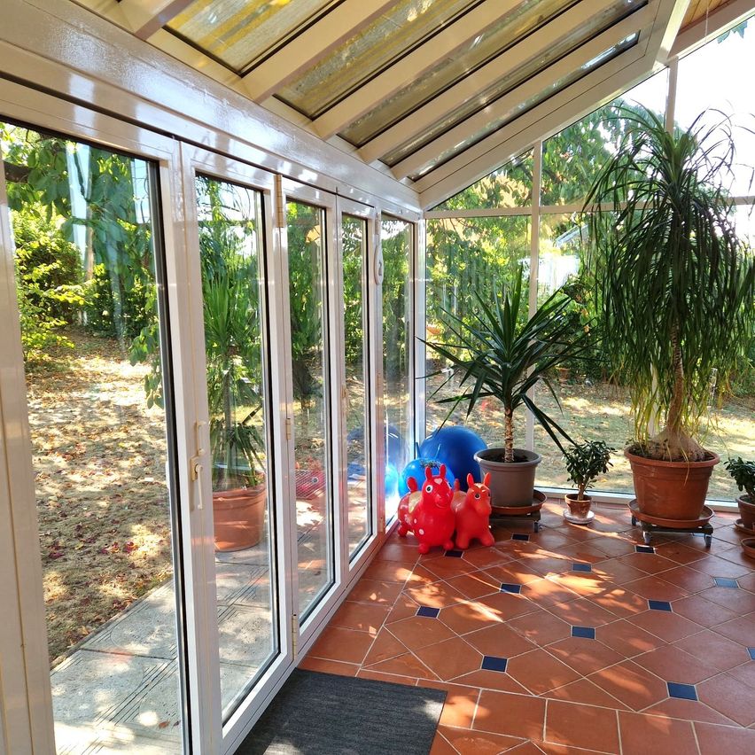 A sunlit conservatory with sliding glass doors, featuring a tiled floor, various potted plants, and two red rabbit toys on the floor.
