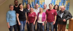 Eine Gruppe von Menschen, sowohl Männer als auch Frauen, posiert für ein Foto in einem Raum mit Holzboden und blauen Vorhängen. Sie lächeln und stehen eng beieinander.