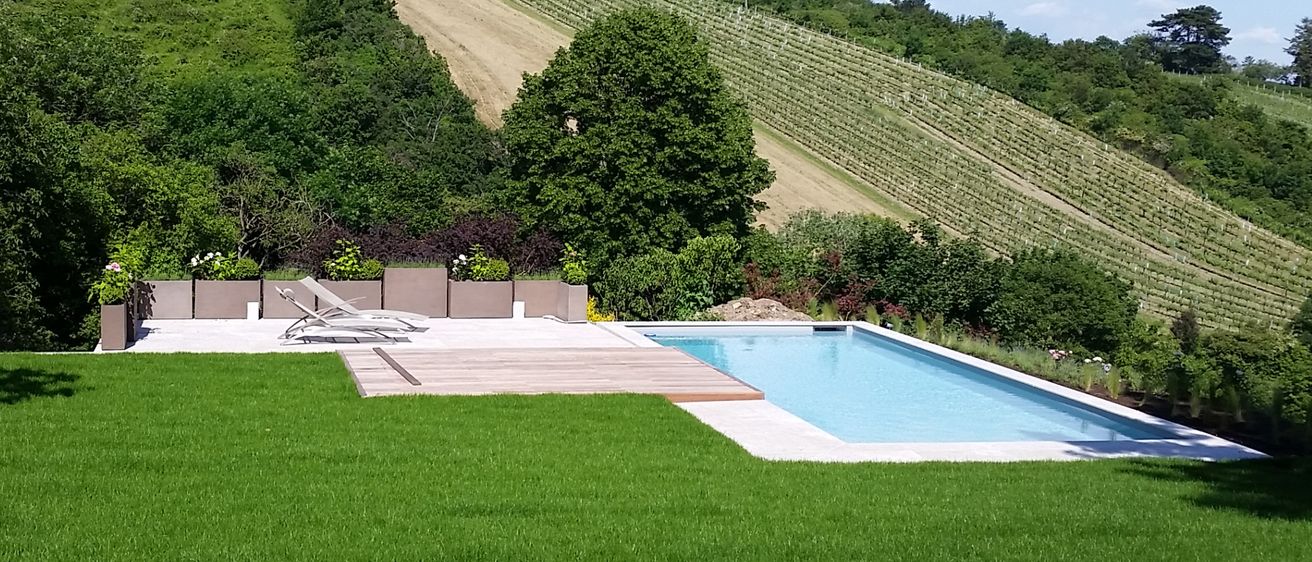 Ein großer Außenpool mit Holzdeck und Liegestühlen, umgeben von üppigem grünem Rasen, Bäumen und einem Weinberg im Hintergrund.