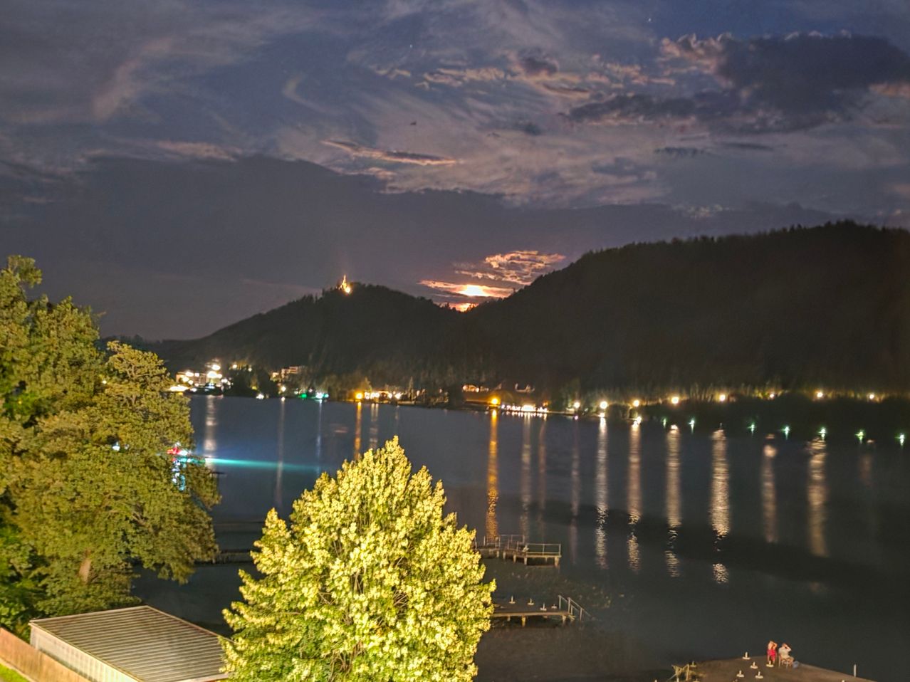 Ein ruhiger Nachtblick auf einen See mit Bergen im Hintergrund, beleuchtet von Stadtlichtern und einer untergehenden Sonne.