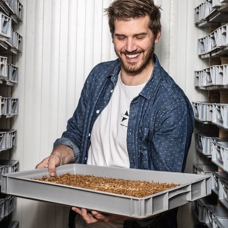 Ein lächelnder Mann hält eine mit braunen Granulaten gefüllte Schale und trägt ein Jeanshemd. Er steht in einem Raum mit Metalllagerboxen.
