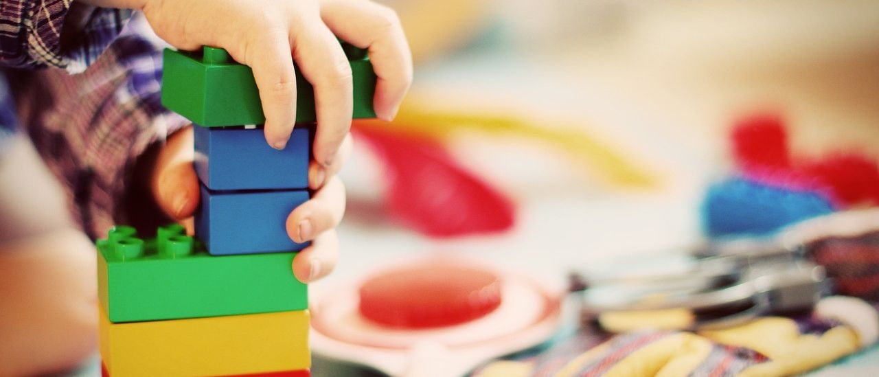 Die Hände eines Kindes stapeln bunte Bauklötze auf einem Tisch mit anderen Spielzeugen im Hintergrund.