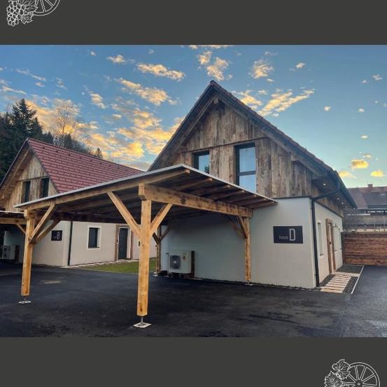Ein Holzhaus mit Carport davor und rotem Dach unter einem blauen Himmel mit Wolken.
