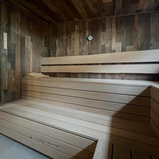 Innenbereich einer Sauna mit Holzbänken und Wänden, einer Uhr an der Wand und einer kleinen Bank auf der unteren Bank.