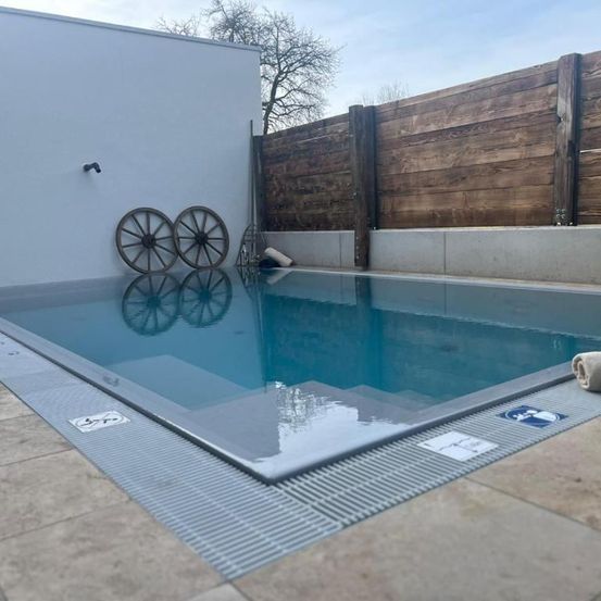 Ein rechteckiger Pool mit grauer Kante und zwei Rädern an der Wand. Der Pool ist leer und hat einen Holzzaun dahinter. Ein Handtuch liegt auf dem Boden.
