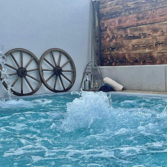 Ein Außenpool mit einer rustikalen Holzwand im Hintergrund. Zwei alte Räder sind an der Wand angebracht. Ein Wasserbrunnen fließt in den Pool. Ein Handtuch ist neben dem Pool platziert.