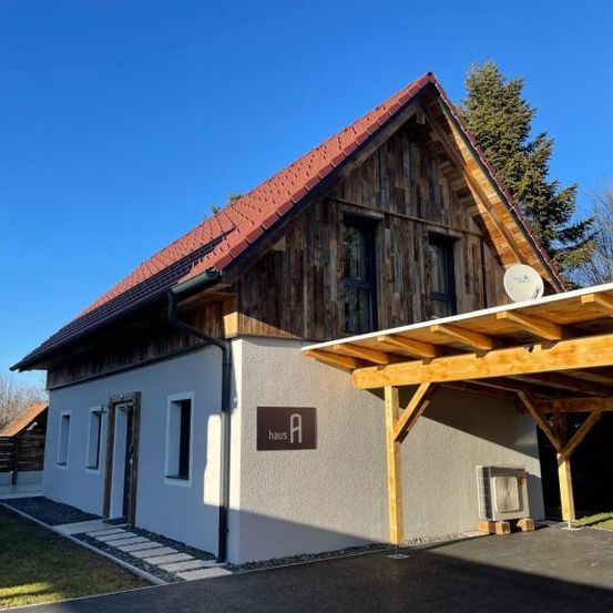 Ein Haus mit einem roten Dach und einer Holzveranda, mit einem Schild, das 'haus A' liest. Es hat einen Schornstein und eine Satellitenschüssel.