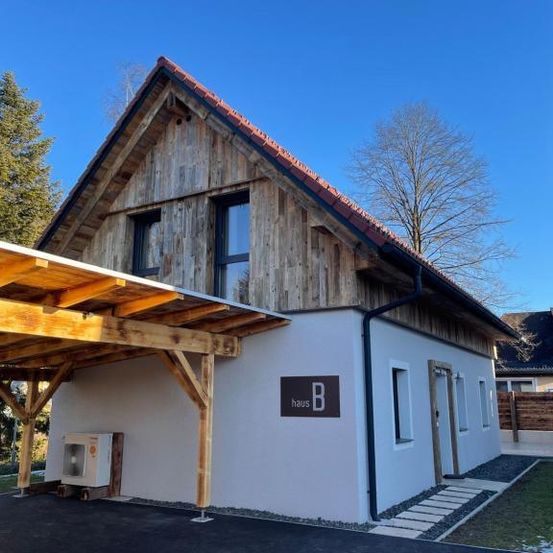 Ein modernes Chalet-Haus mit Holzfassade, rotem Dach und weißen Wänden. Es hat ein Carport, Fenster und ein Schild 'Haus B'.