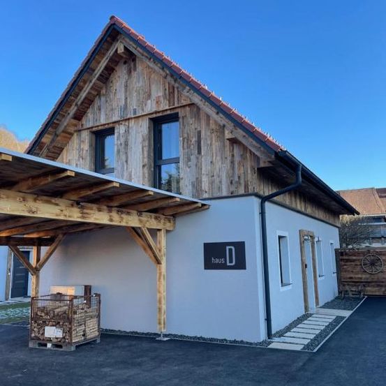 Ein Holzhaus mit rotem Dach und überdachtem Eingang steht auf einer Einfahrt. In der Nähe des Eingangs befindet sich ein Stapel Holz.