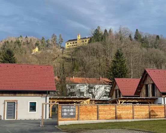 Eine kleine Stadt mit Häusern und einem Gebäude auf einem Hügel unter einem bewölkten Himmel. Ein Holzzaun und ein Rasen davor.