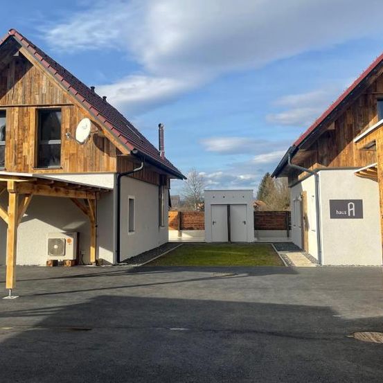 Zwei Holzhäuser mit Carport. Ein Schild auf dem Haus steht 'Haus A'. Der Himmel ist bewölkt.