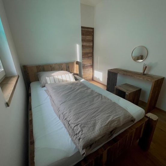 Ein kleines Schlafzimmer mit einem Holzbett, einem runden Spiegel, einem Schreibtisch und einer Holztür. Der Raum ist von Sonnenlicht durchflutet.