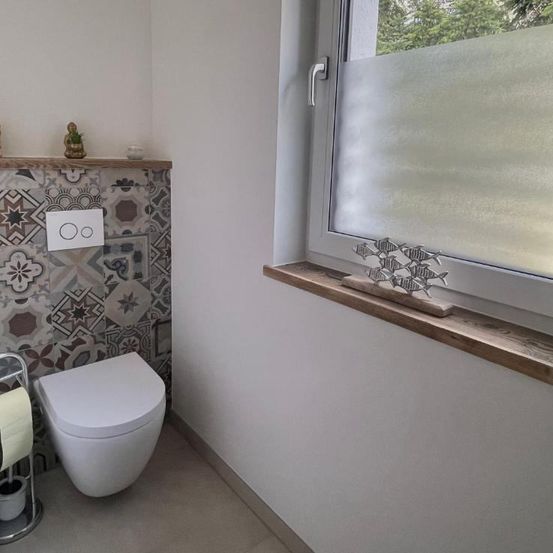 Ein kleines Badezimmer mit einer weißen Toilette, einem wandmontierten Schalter und einer gefliesten Rückwand. Rechts ist ein Fenster mit mattiertem Glas zu sehen.