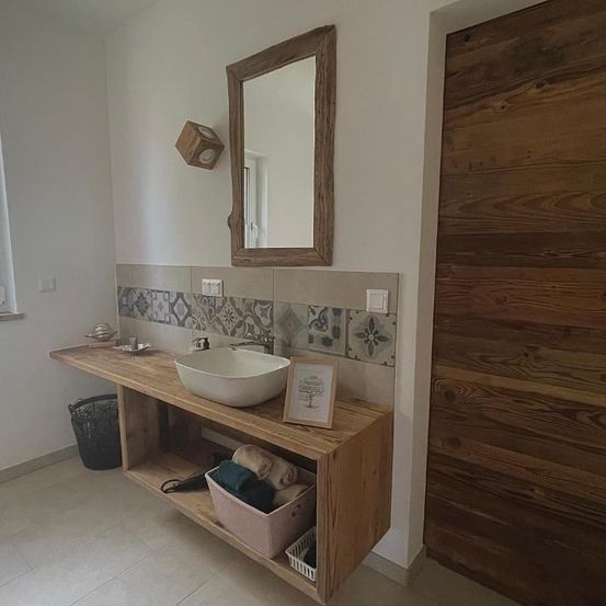 Ein Badezimmer mit einem hölzernen Waschtisch hat ein Waschbecken, einen Spiegel und eine Holztür. Ein Bilderrahmen ist an der Wand montiert.