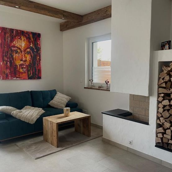 Ein Wohnzimmer mit einer blauen Couch, einem Holztisch und einem Kamin. Die Wand hat ein großes Gemälde eines Frauengesichts.